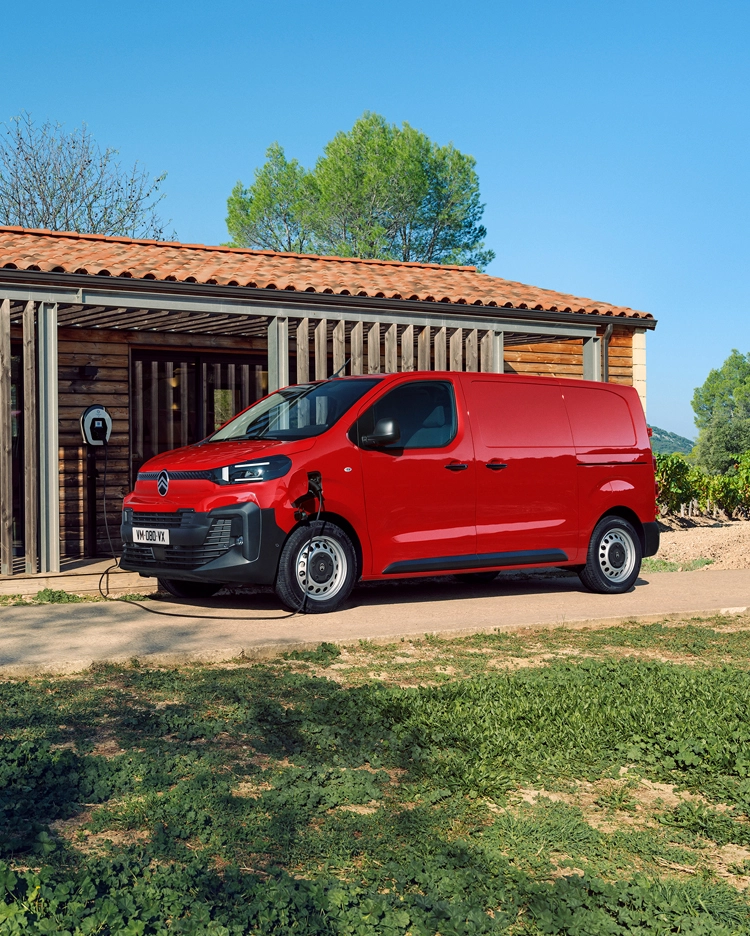 Side three‑quarter exterior view of a red Citroën ë‑Jumpy charging in front of a building, showing modern electric design, everyday practicality, connectivity and confident professional presence.